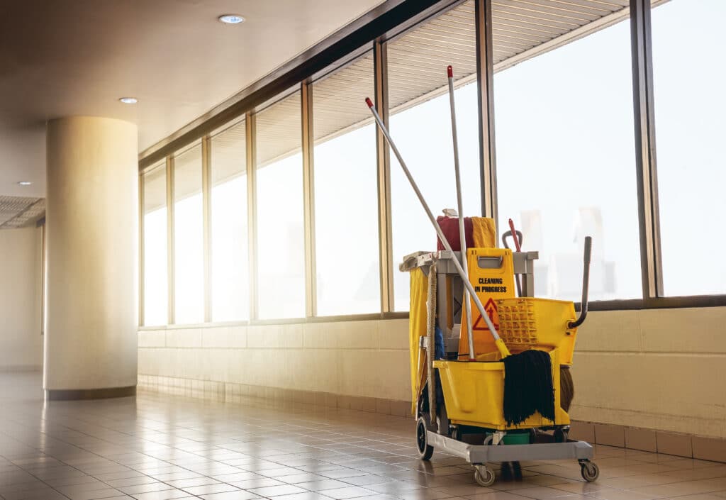 The image captures a bustling scene in a hallway where cleaning supplies are in action. At the center of the frame stands a yellow cart brimming with various cleaning products