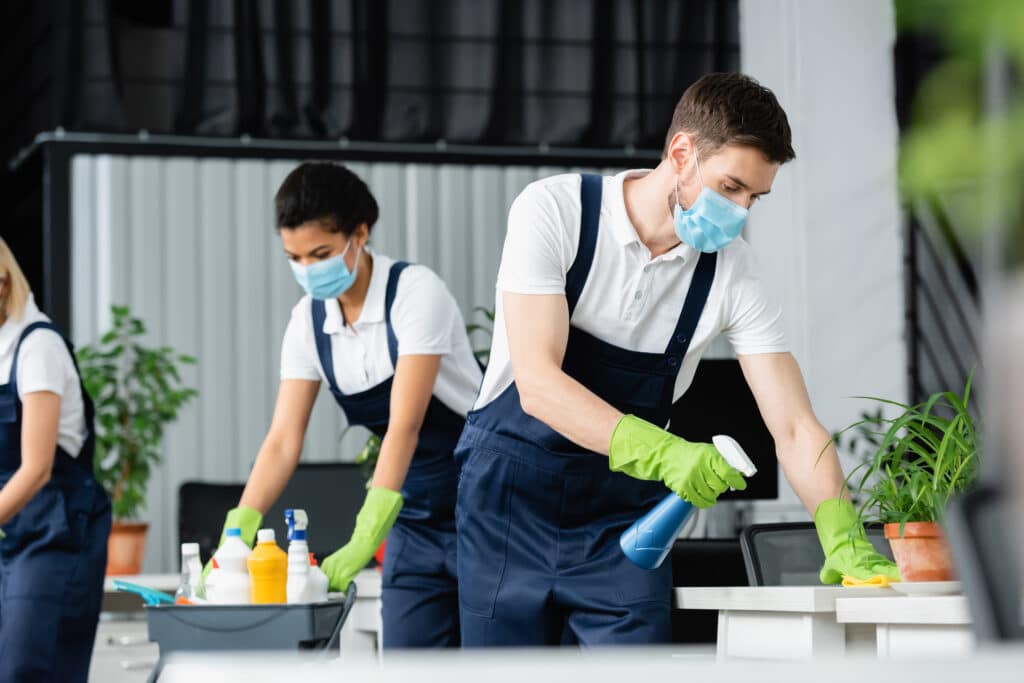 A group of people is cleaning an office. A woman wears overalls, a mask, and gloves. A man is in a white shirt and a surgical mask. Another person has green gloves and holds a blue spray bottle.