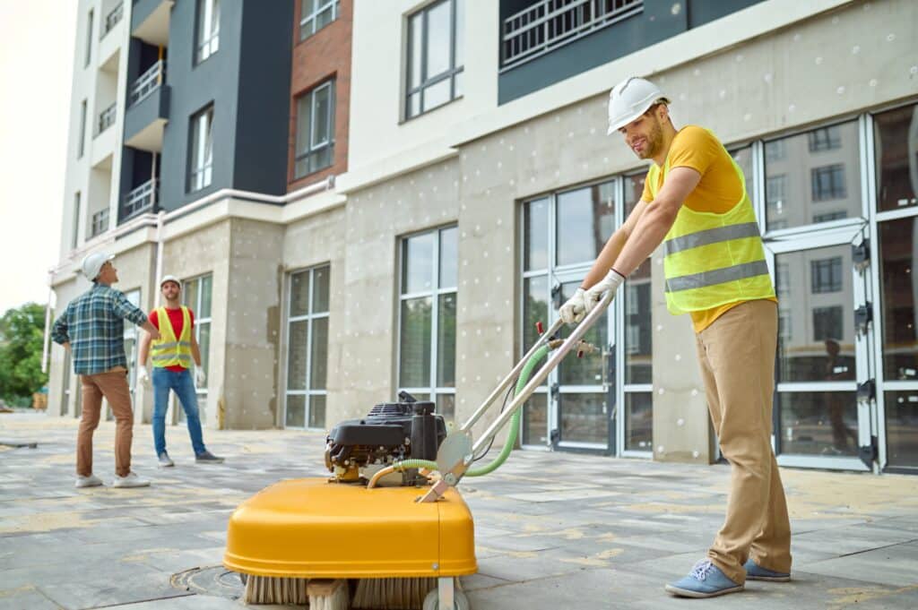 Construction workers sweep, grind, and excavate on a bustling site. Two workers stand by a building. One holds a shovel, and the other is on his phone. In the background, a group gathers to work together