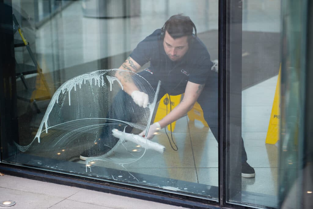 A corporate cleaner wearing headphones carefully wipes a glass door with a cloth. A yellow sign stands on the sidewalk, adding interest to the scene.